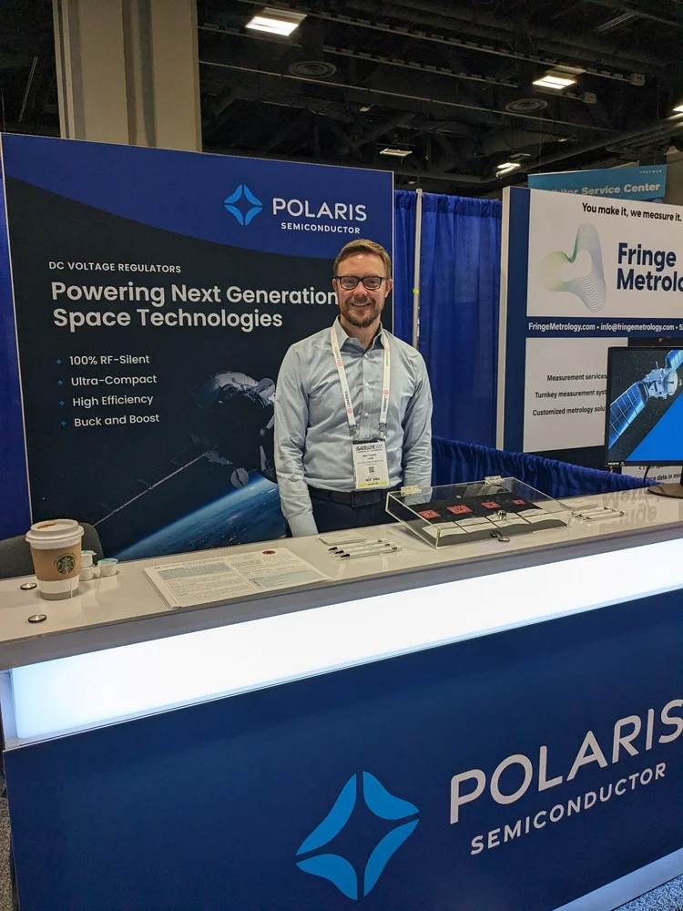 Dr. Matthew Lumb standing at the Polaris Semiconductor conference booth, with a banner reading 'DC voltage regulators — powering next generation space technologies' and sample chips on the counter.