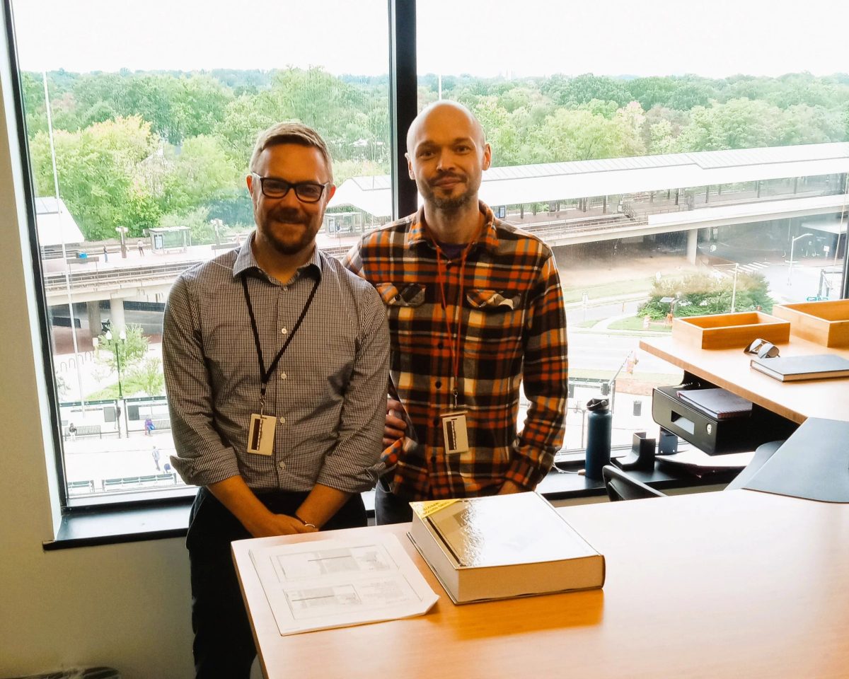 Matthew Lumb and Kenneth Schmieder standing side by side at an office desk with a printed schematic and a thick reference book, in front of large windows overlooking an elevated rail station.