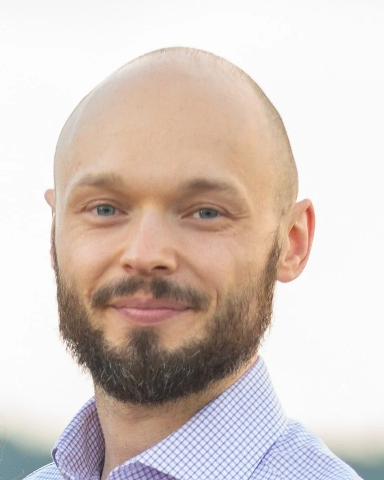 Portrait of Dr. Kenneth Schmieder, smiling, with a shaved head and full beard, wearing a light checked button-down shirt against a bright background.
