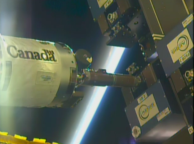 Exterior view from the International Space Station showing a robotic arm marked 'Canada' maneuvering hardware beside externally mounted experiment payloads, with Earth's atmosphere glowing along the horizon.